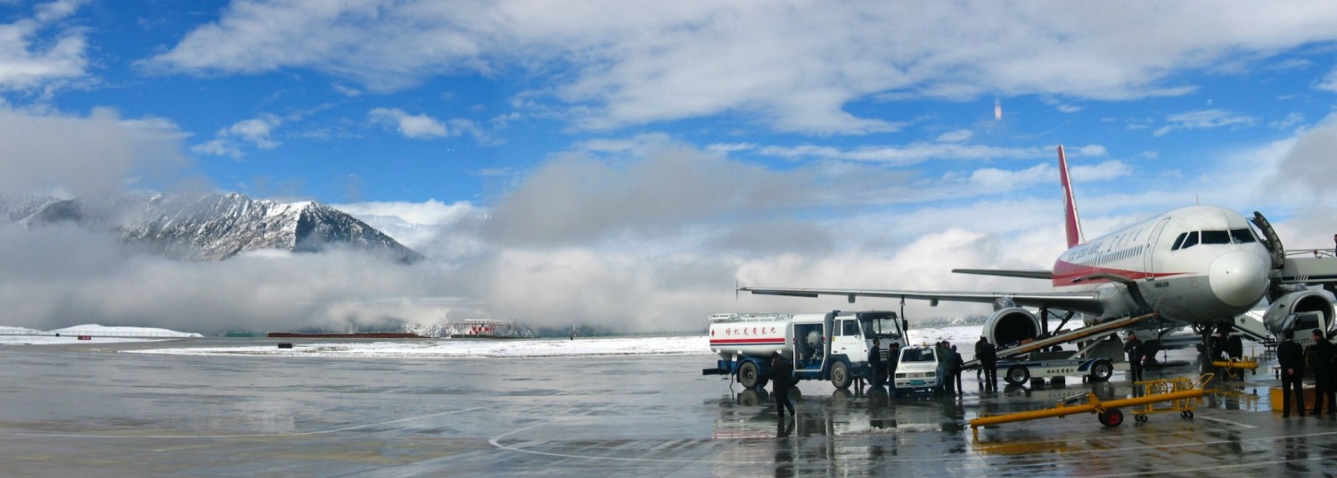  A snowy runway with a plane and vehicles on it, with a snow-capped mountain in the background.